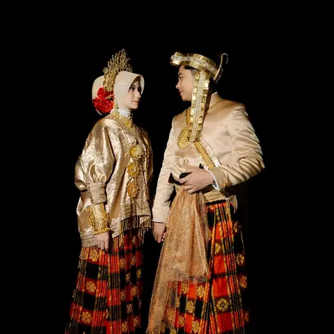 Cantika and Deni in gold-embroidered jackets and woven red-and-gold sarongs, facing each other against black
