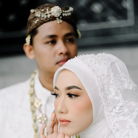 Fadli and Aini on their wedding day: the bride in a white hijab and jasmine strands, her hand at her cheek, the groom behind her in white with a beaded headdress
