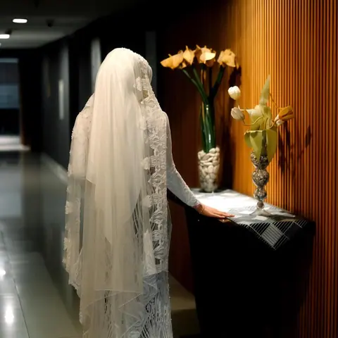 Aini, the bride, in a long lace veil and gown, at a side table of flowers in a hotel corridor