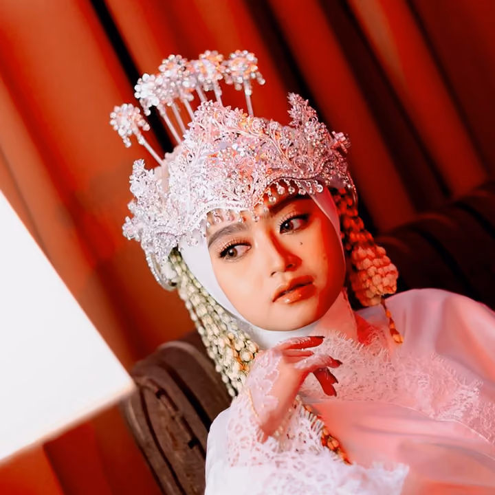 The bride of the Syahla and Bagas wedding in a pink-and-white beaded headdress, her chin on her hand, against red curtains