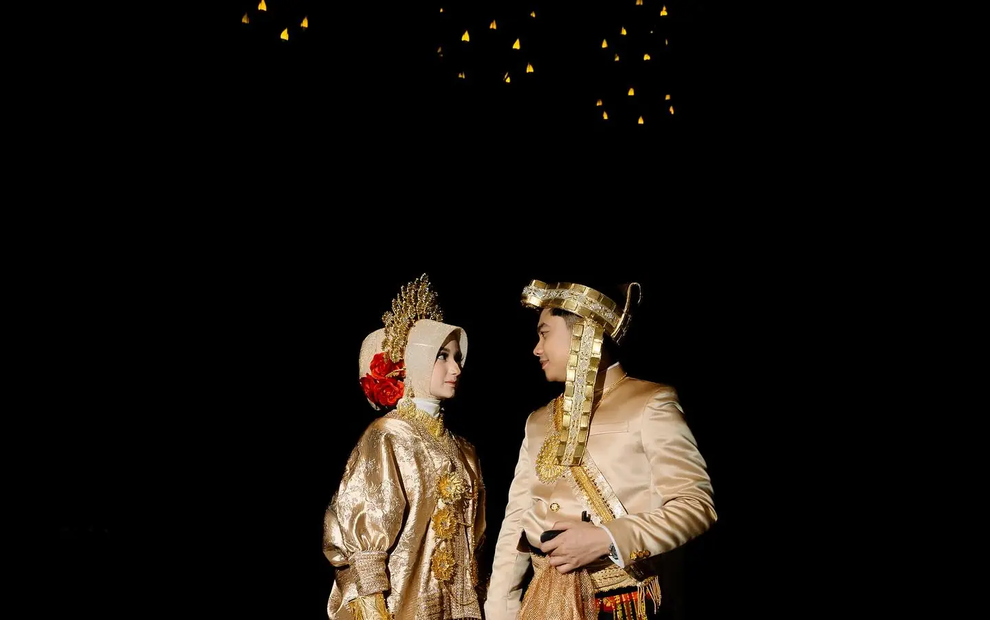 Cantika and Deni on their wedding day, in gold-embroidered jackets and woven red-and-gold sarongs, facing each other under small gold lights against black