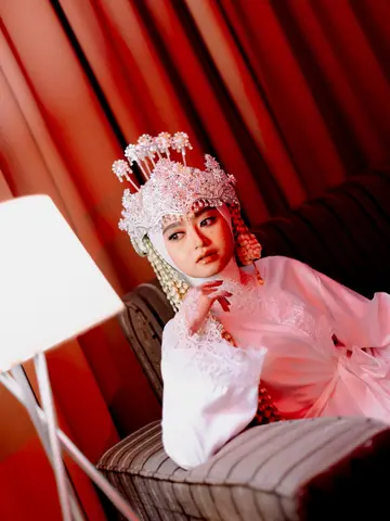 A bride in a pink-and-white beaded headdress and pink dress, her chin on her hand, against red curtains