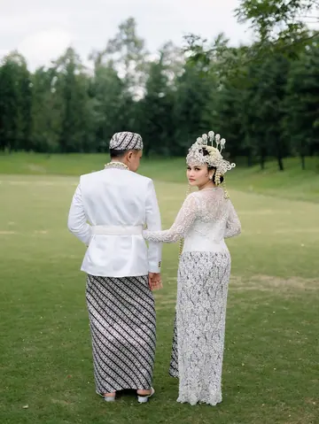 A bride and groom seen from behind on a lawn, she in a lace kebaya and crown, he in white with a batik sarong