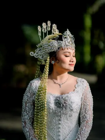 A bride in a silver crown with long jasmine strands and a lace kebaya, eyes closed, against dark leaves